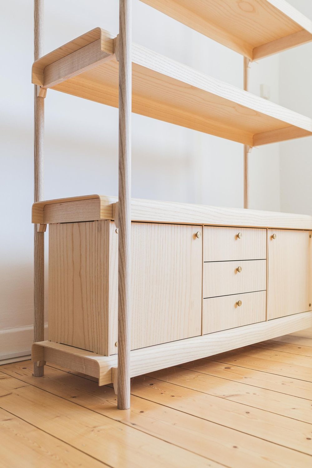 Ein minimalistisches Holzregal mit drei offenen Regalen und einem Schrank mit Schubladen, in hellen Holztönen und mit Messinggriffen.