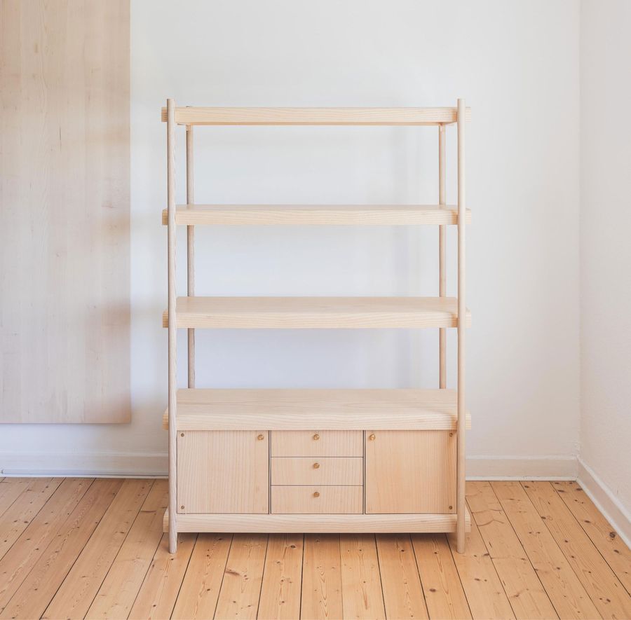 Ein minimalistisches Holzregal mit vier offenen Regalen und einem Schrank unten, vor einer hellen Wand und einem Holzboden.