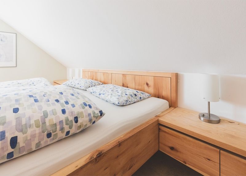 Gemütliches Schlafzimmer mit einem Holzbett mit gemusterter Bettwäsche, einem Nachttisch und einer modernen Lampe vor einer Dachschräge.