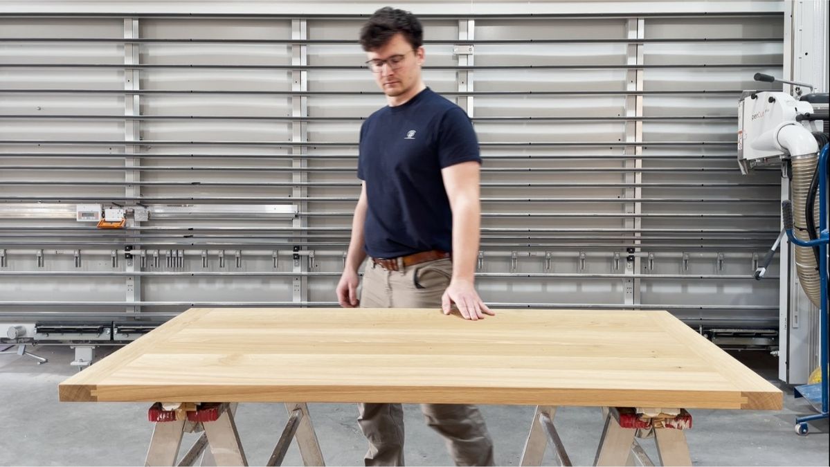 Felix Zintel steht in einer Werkstatt neben einem Holztisch auf Sägeböcken. Im Hintergrund sind Werkzeuge und Geräte zu sehen.