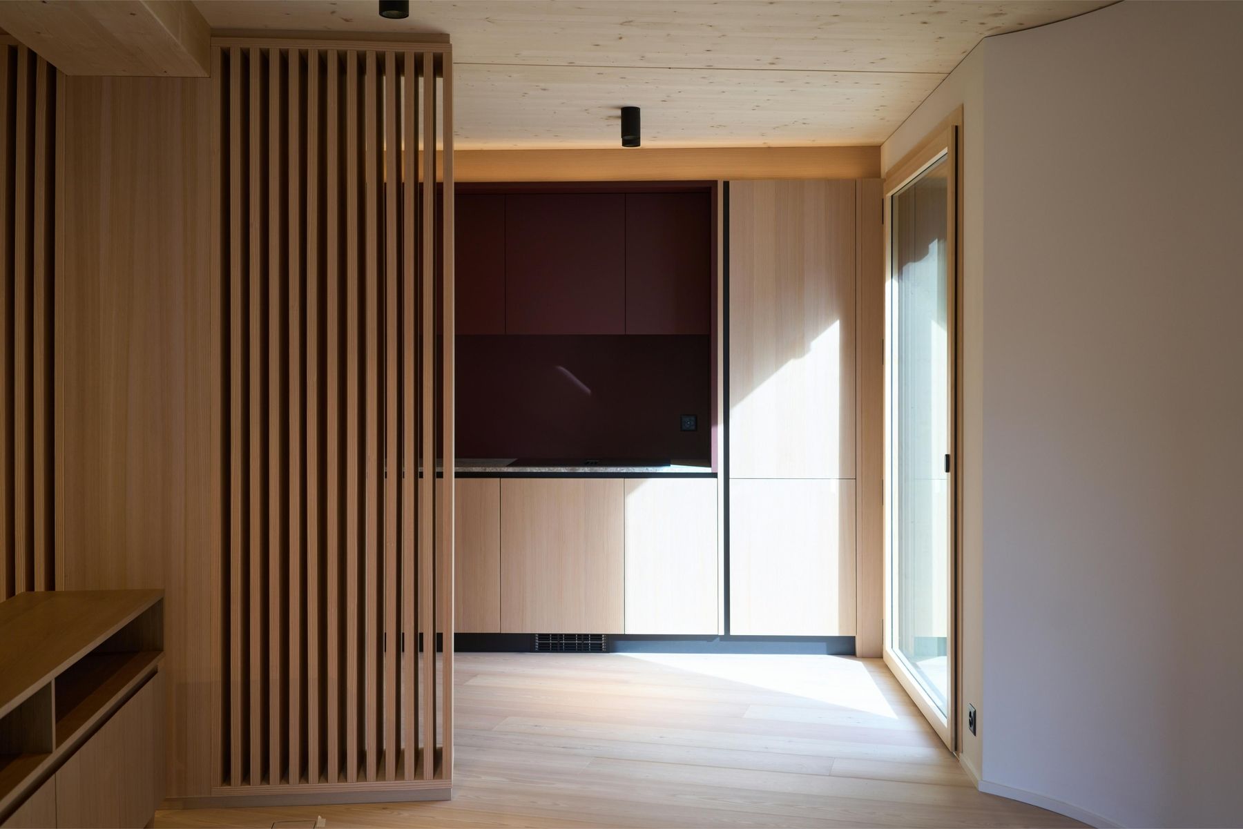 Minimalistisches Interieur mit Holzvertäfelung, einer kompakten Küche mit burgunderfarbenen Akzenten und reichlich natürlichem Licht durch ein Fenster.