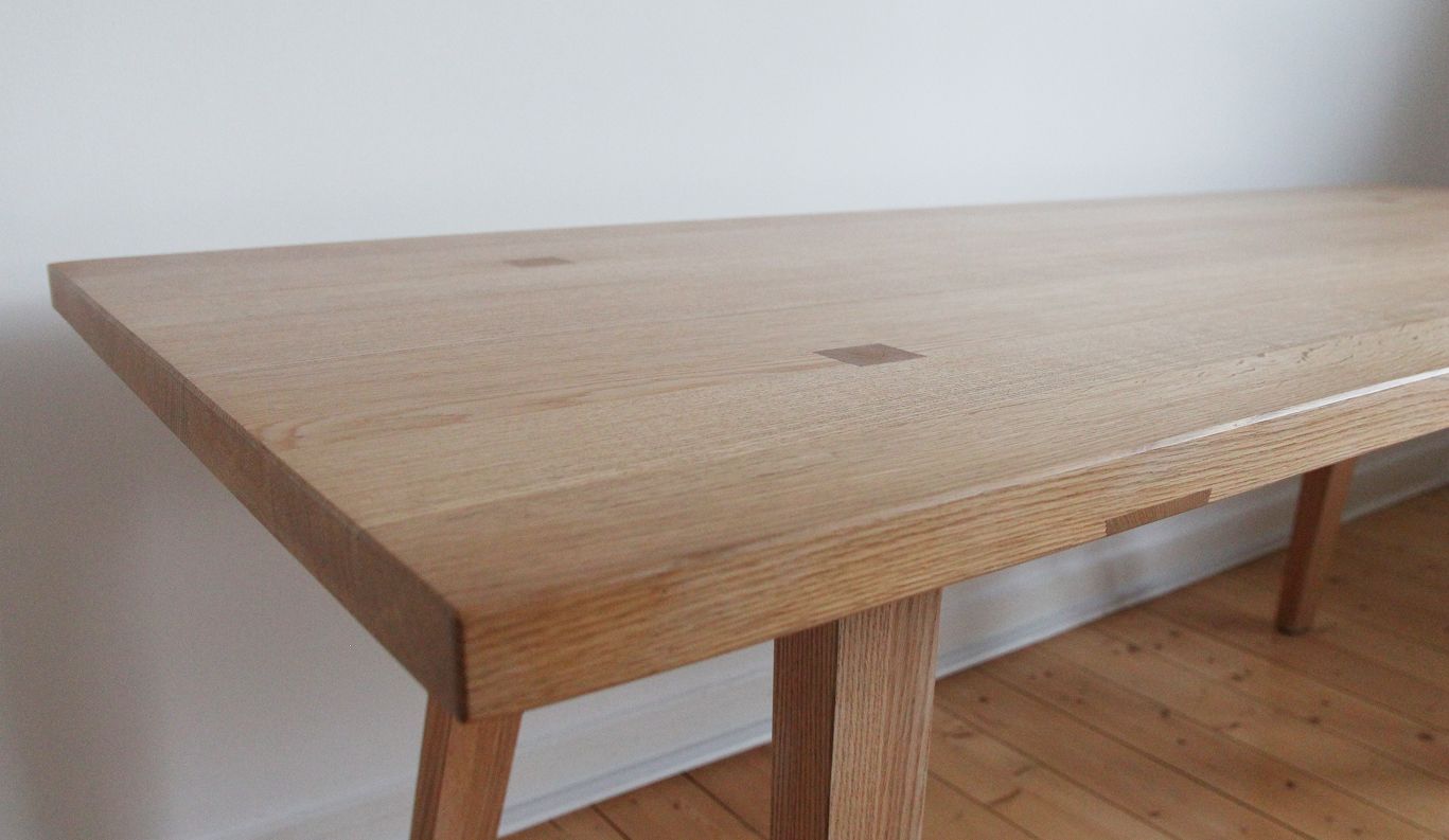 Nahaufnahme eines Holztisches mit glatter, heller Eichenoberfläche und quadratischen Intarsiendetails vor einer schlichten, neutralen Wand.